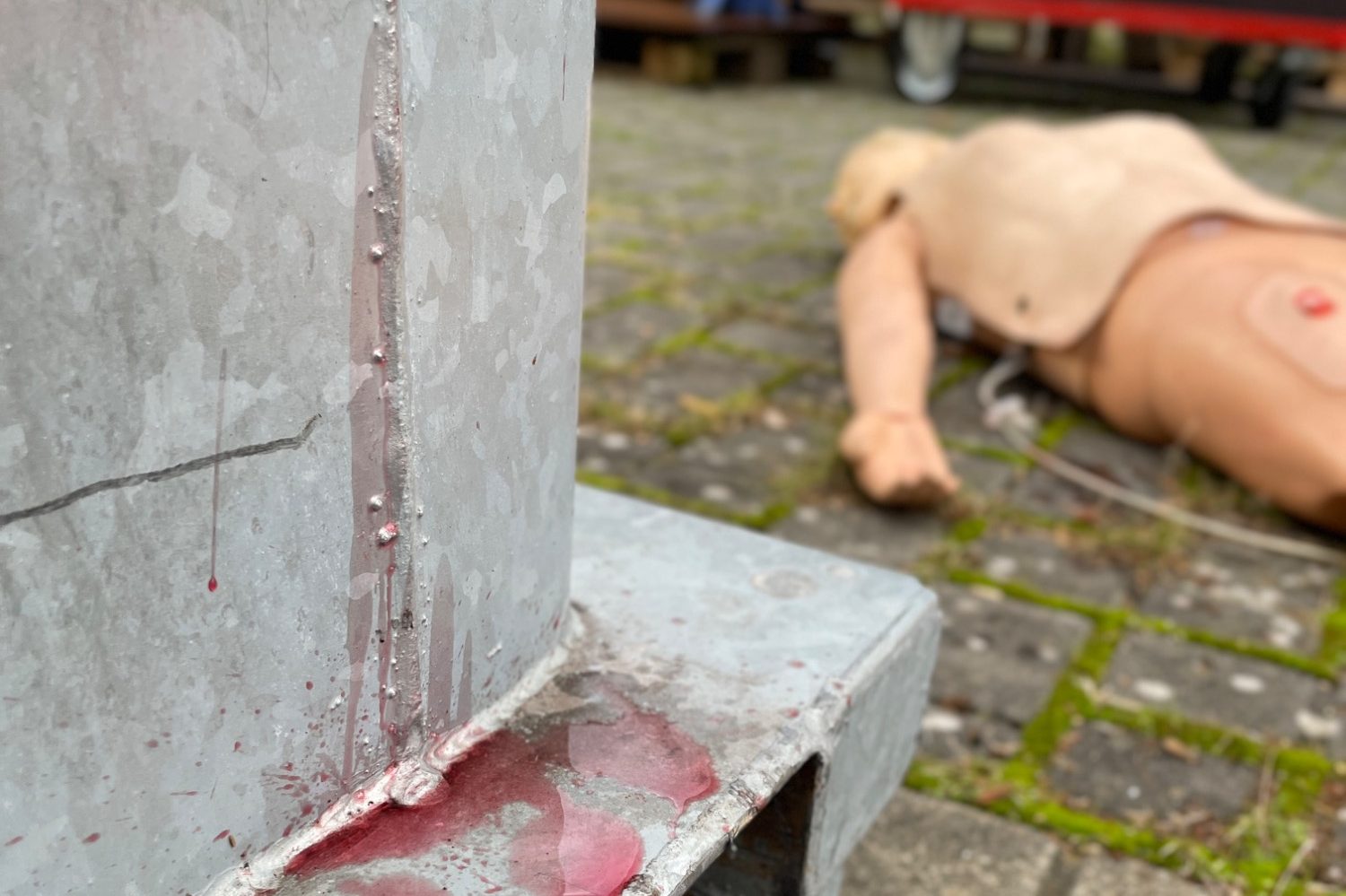 Ein Dummy im liegt im Hintergrund auf dem Boden. Im Vordergrund sieht man Blutanhaftungen an einem metallenem Objekt