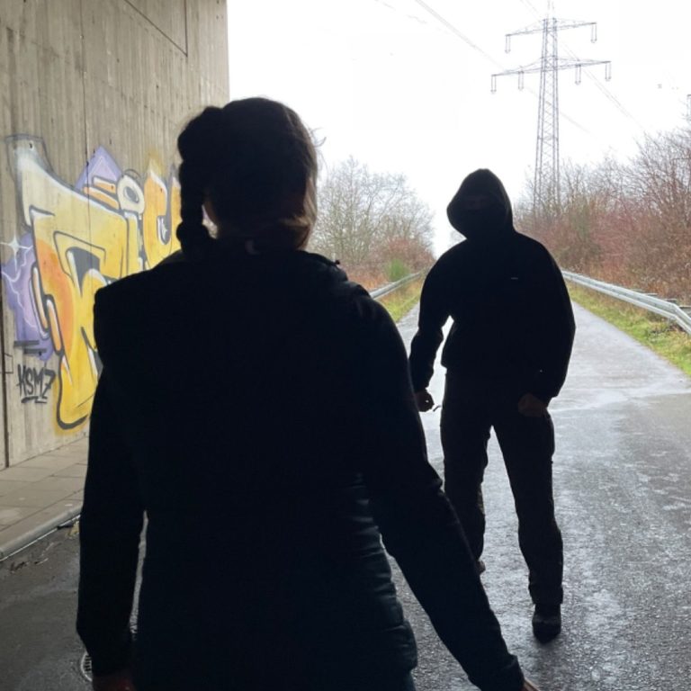 Deeskalation und Selbstverteidigung für Frauen Eine Frau steht in einer dunklen Gasse und wird von einem vermummten Mann bedroht