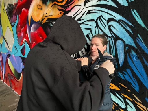Eine Frau wird mit einem Messer Bedroht und an eine Wand mit einem Graffiti gedrückt