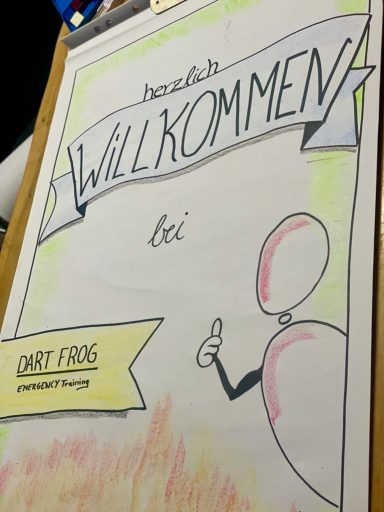 Eine Flipchart mit der Aufschrift "Herzlich Willkommen bei Dartfrog Emergency Training" Eine gezeichnete Figur zeigt Daumen hoch