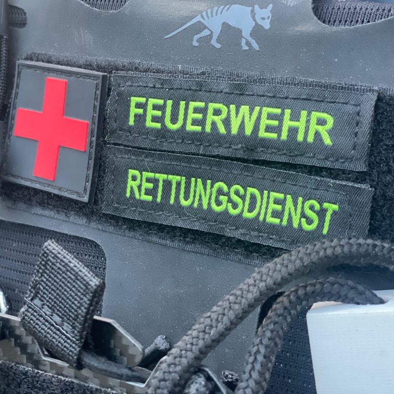 Ein Plattenträger mit der Aufschrift Feuerwehr unf Rettungsdienst. Daneben ist ein Patch mit einem Rot-schwarzen Kreuz